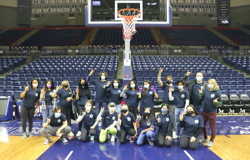 A group of students gather for a picture on a basketball court. They each hold up the UConn hand signal. 